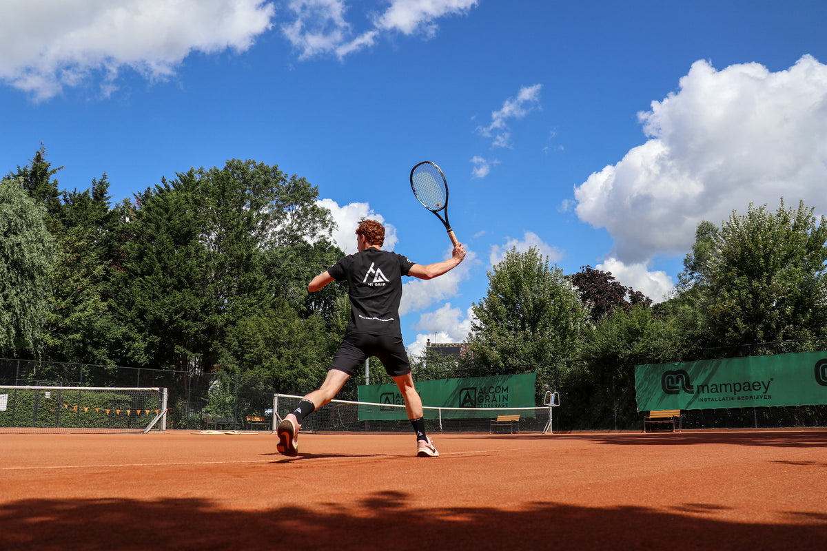 Tennisser in een donkerblauw shirt serveert op een gravelbaan, met witte HÏ Grip-gripsokken in zijn tennisschoenen