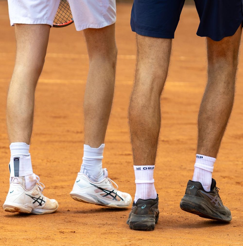 Benen van twee tennissers op gravel, een van hen draagt witte HÏ Grip-gripsokken in zijn tennisschoenen